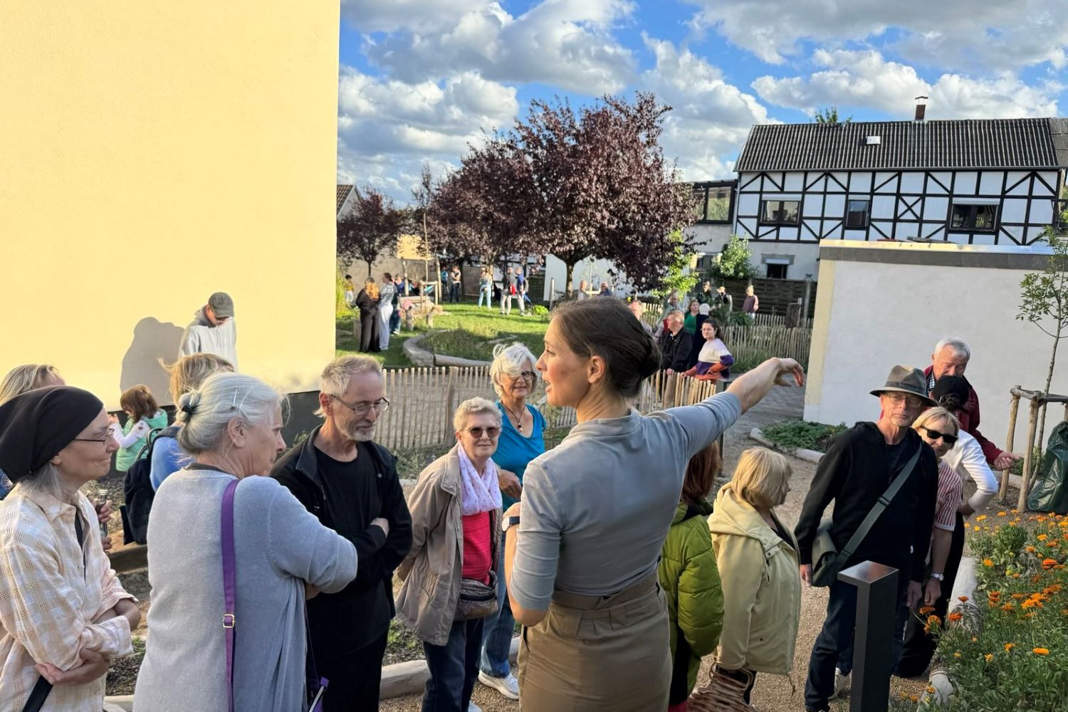 Viele Leute auf der PIkoParkfläche hören der Naturgartenplanerin bei ihren Erläuterungen zu. Die Sonne scheint und die ersten Beete blühen in gelb und orangenen Farben