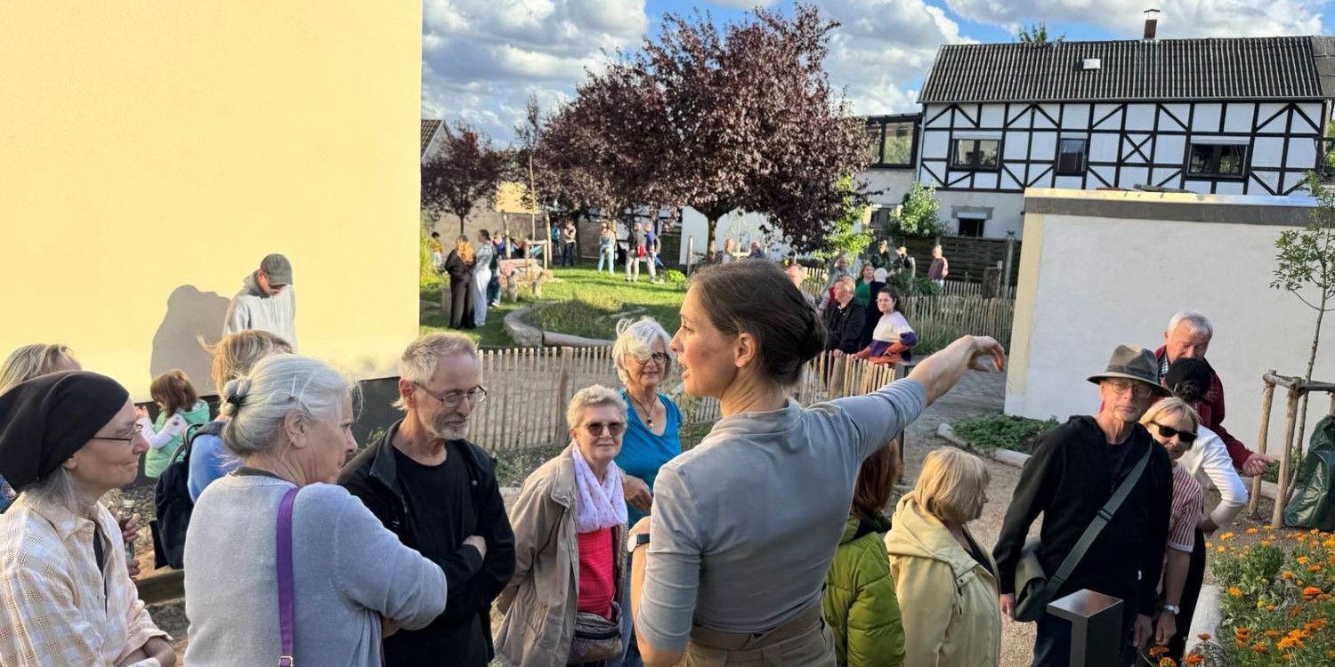 Viele Leute auf der PIkoParkfläche hören der Naturgartenplanerin bei ihren Erläuterungen zu. Die Sonne scheint und die ersten Beete blühen in gelb und orangenen Farben
