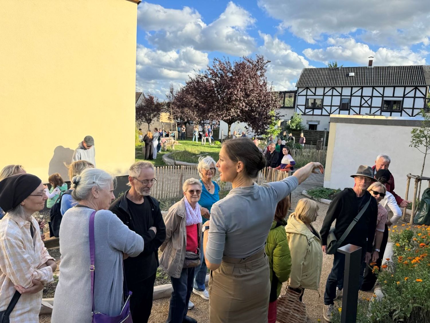 Viele Leute auf der PIkoParkfläche hören der Naturgartenplanerin bei ihren Erläuterungen zu. Die Sonne scheint und die ersten Beete blühen in gelb und orangenen Farben