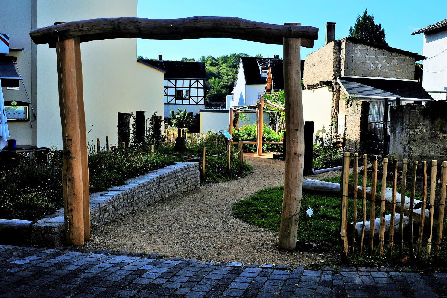 Eingang und Überblick über die Fläche vom fertig gestellten PikoPark in Sinzig. Ein Eingangstor aus Baumstämmen. Blick auf wassergebundene Wegedecke eine Trockenmauer Hochbeet