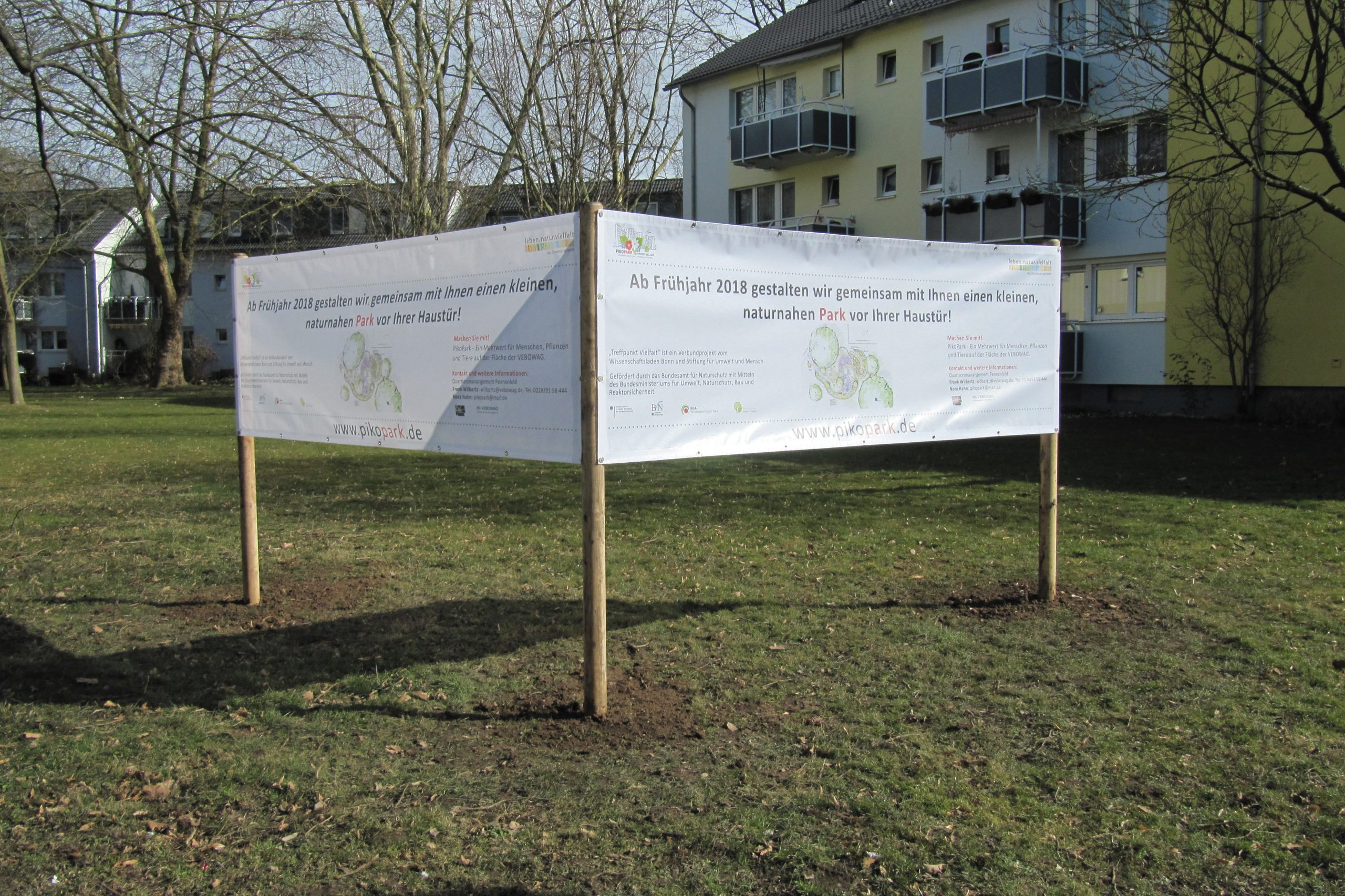 Zwei übers Ecke aufgestellte große Banner auf der Wiese neben Wohnriegel. Die Bauschilder informieren Vorbeigehende über das Bauvorhaben des PikoParks