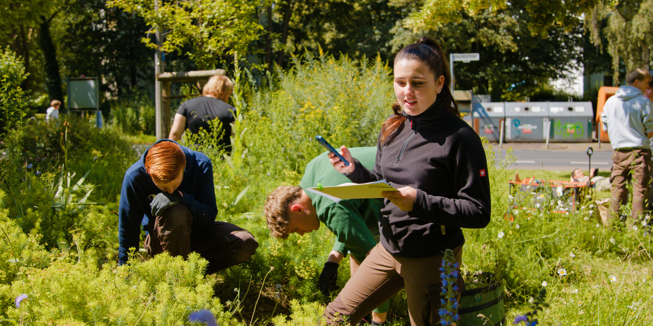 Eine junge Frau steht an einem Blumenbeet mit Unterlagen in der einen und ihrem Smartphone in der anderen Hand und schaut auf die Pflanzen. Neben ihr jäten zwei junge Männer.