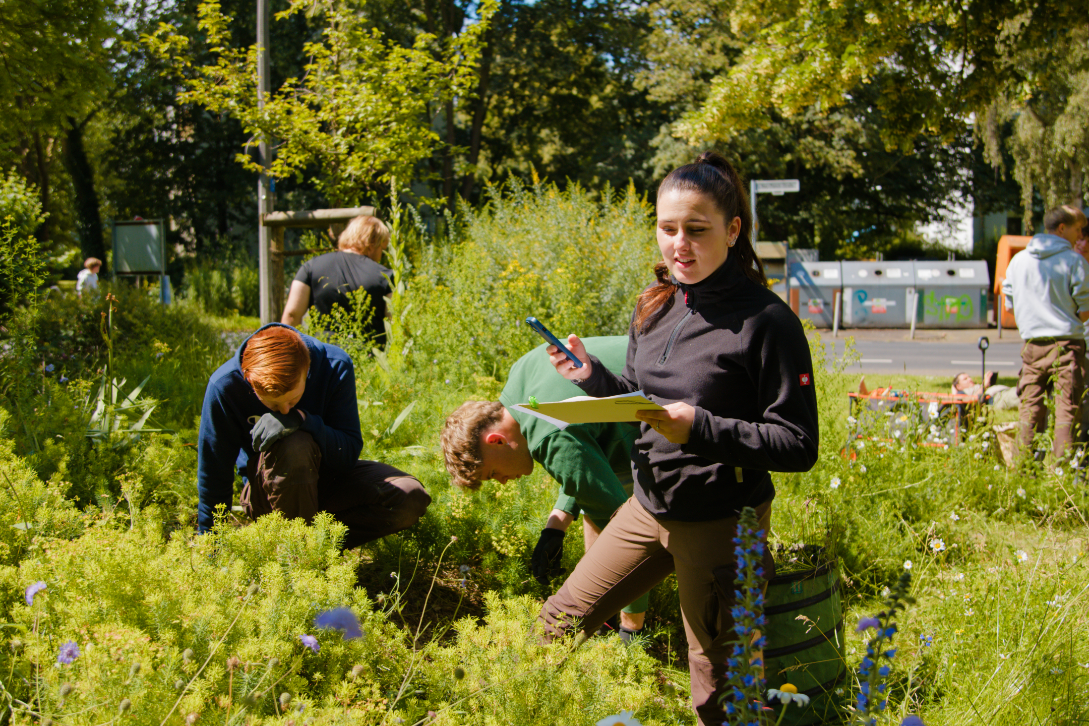 Eine junge Frau steht an einem Blumenbeet mit Unterlagen in der einen und ihrem Smartphone in der anderen Hand und schaut auf die Pflanzen. Neben ihr jäten zwei junge Männer.