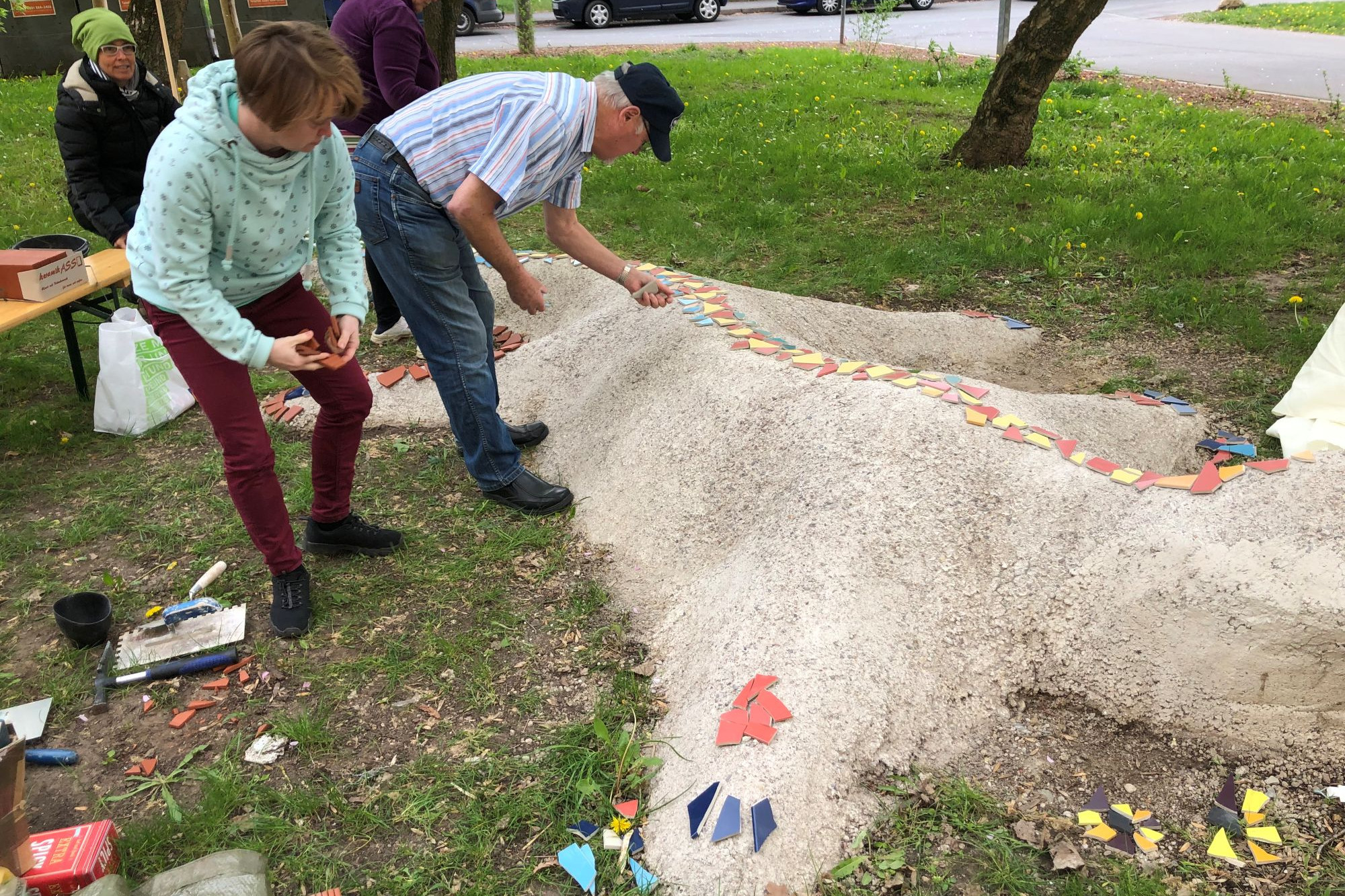 Eine Frau und ein Mann verzieren eine Salamander Betonskulptur, die auf einer Wiese liegt mit Mosaiksteinen