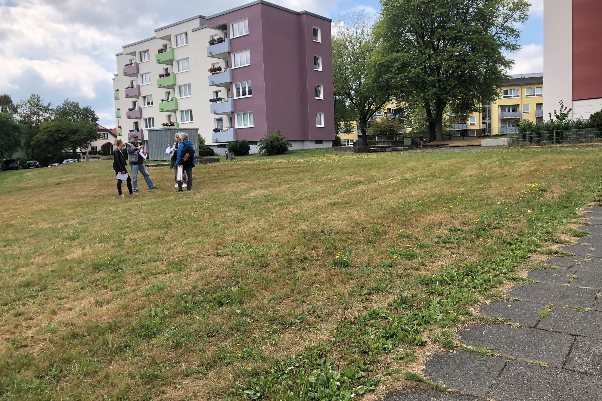 Vier Erwachsene stehen auf einer größeren Rasenfläche. Im Hintergrund 4-schossige Wohnblöcke
