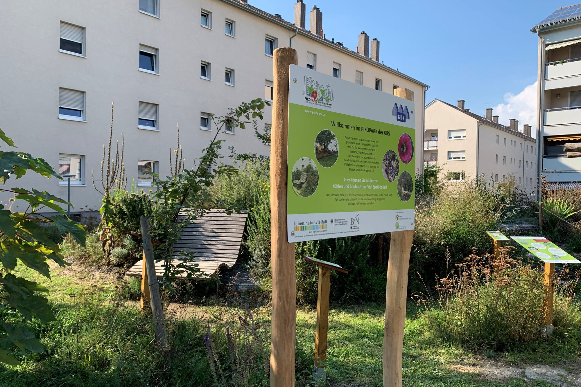 Blick auf den PikoPark in Speyer. Im Vordergrund ein grünes Willkommensschild mit Infos zum Park. Im HIntergrund de Park mit einer geschwungenen Holzbank und weiter hinten 2 Wohnriegel mit 4 Etagen