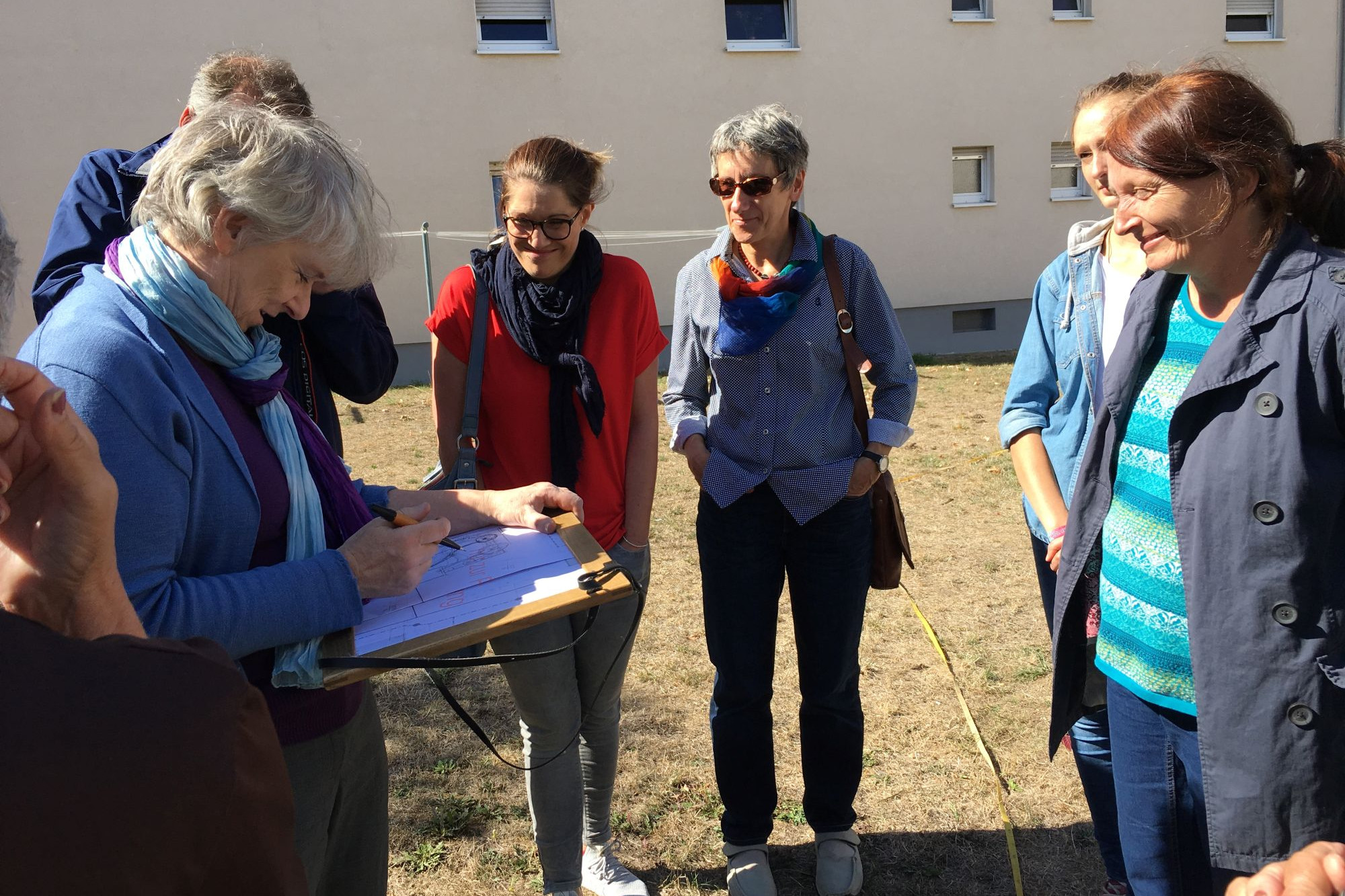Eine Frau skizziert auf einer vertrockneten Wiese auf einem Klemmbrett. Um sie herum stehen 6 Erwachsene und schauen, was sie skizziert. Im Hintergrund ist eine Wohngebäude zu sehen.