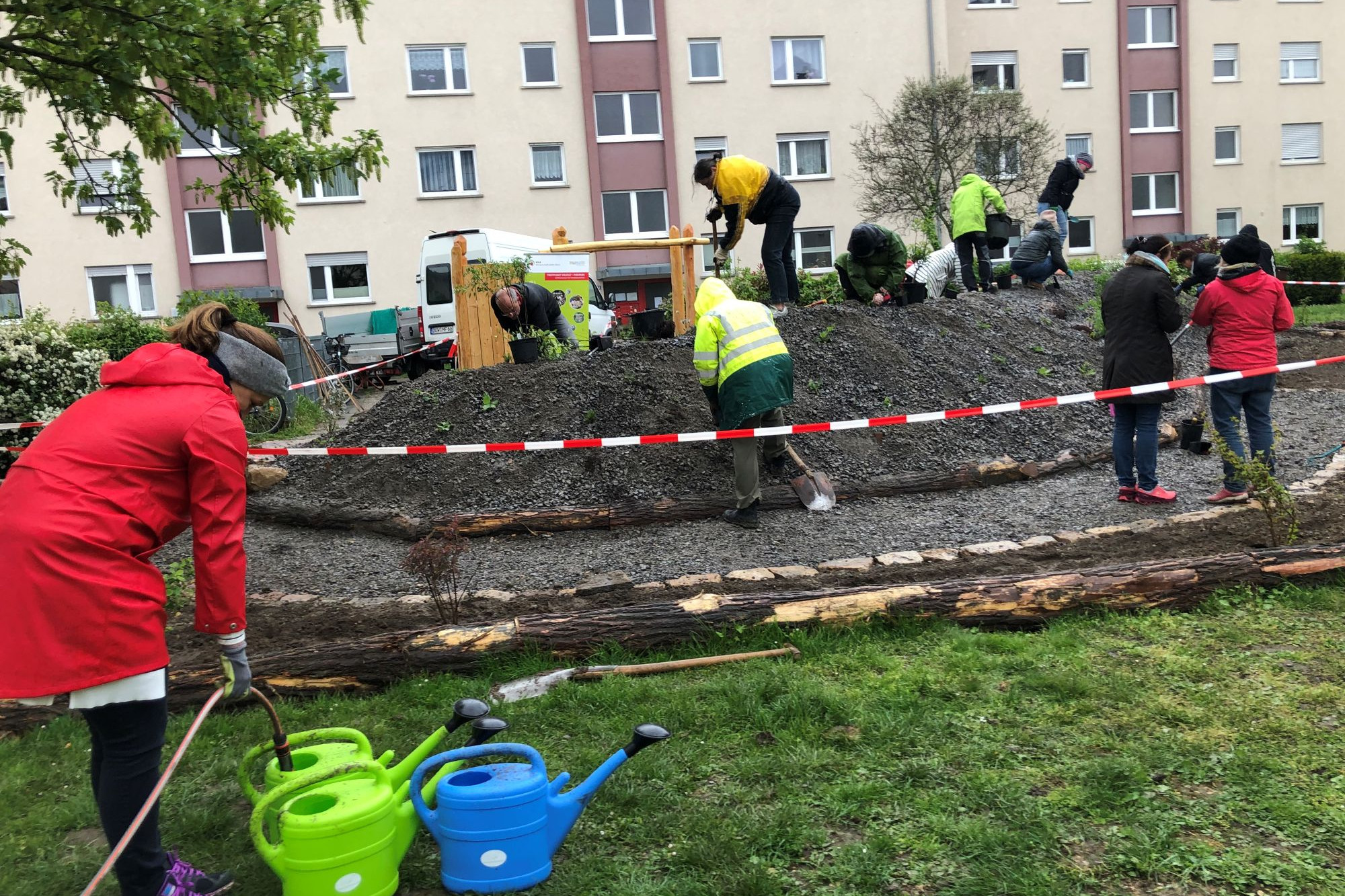 Zwölf Männer und Frauen pflanzen Stauden auf einem Schotterhügel . Im Vordergrund befüllt eine Frau im roten Regenmantel 3 grüne und blaue Gießkannen.