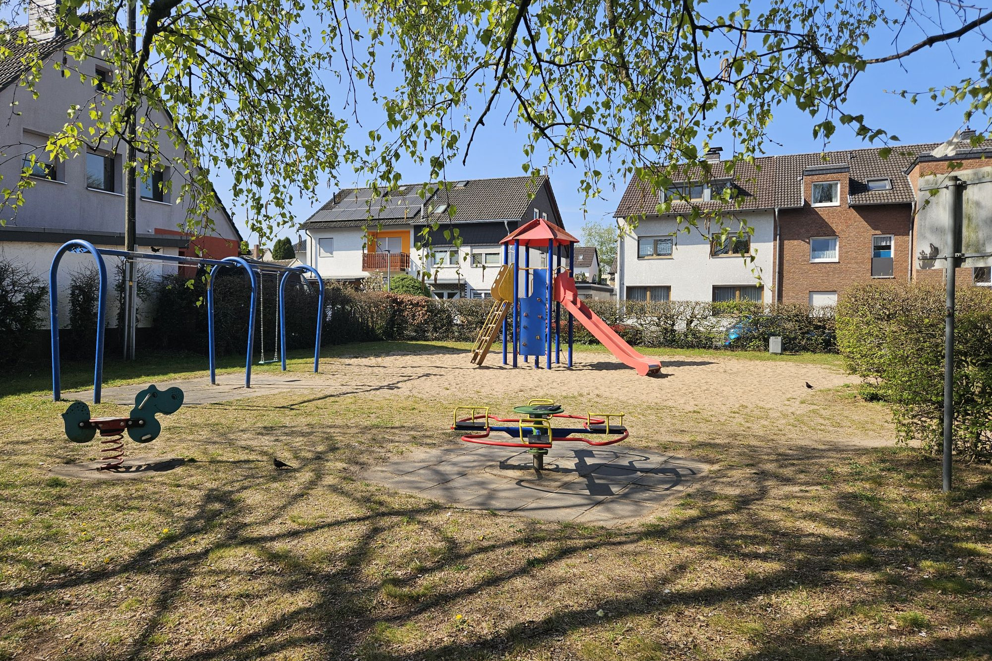 Vlick auf einen alten Speilplatz auf einer sandigen Fläche. Eine rot-blaue Rutsche und ein kleines Drehkarussel. Hinter der einrahmenden Hecke sind Mehrfamilienhäuser zu erkennen