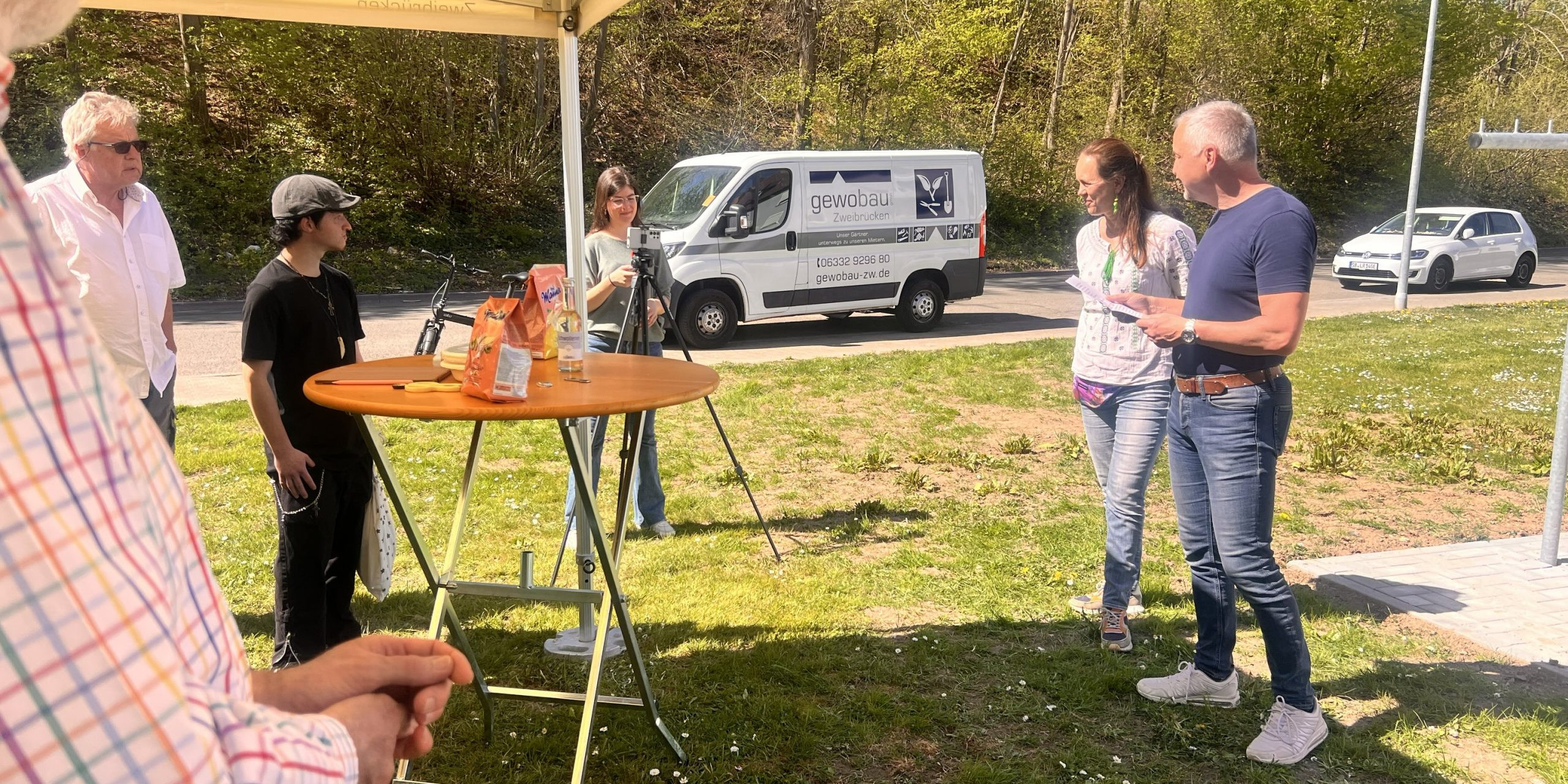 Infoveranstaltung auf einer Rasenfläche. Ein Redner im blauen T-Shirt. Eine Frau in weißer Bluse steht daneben. An einem Stehtisch sieht man Zuhörer