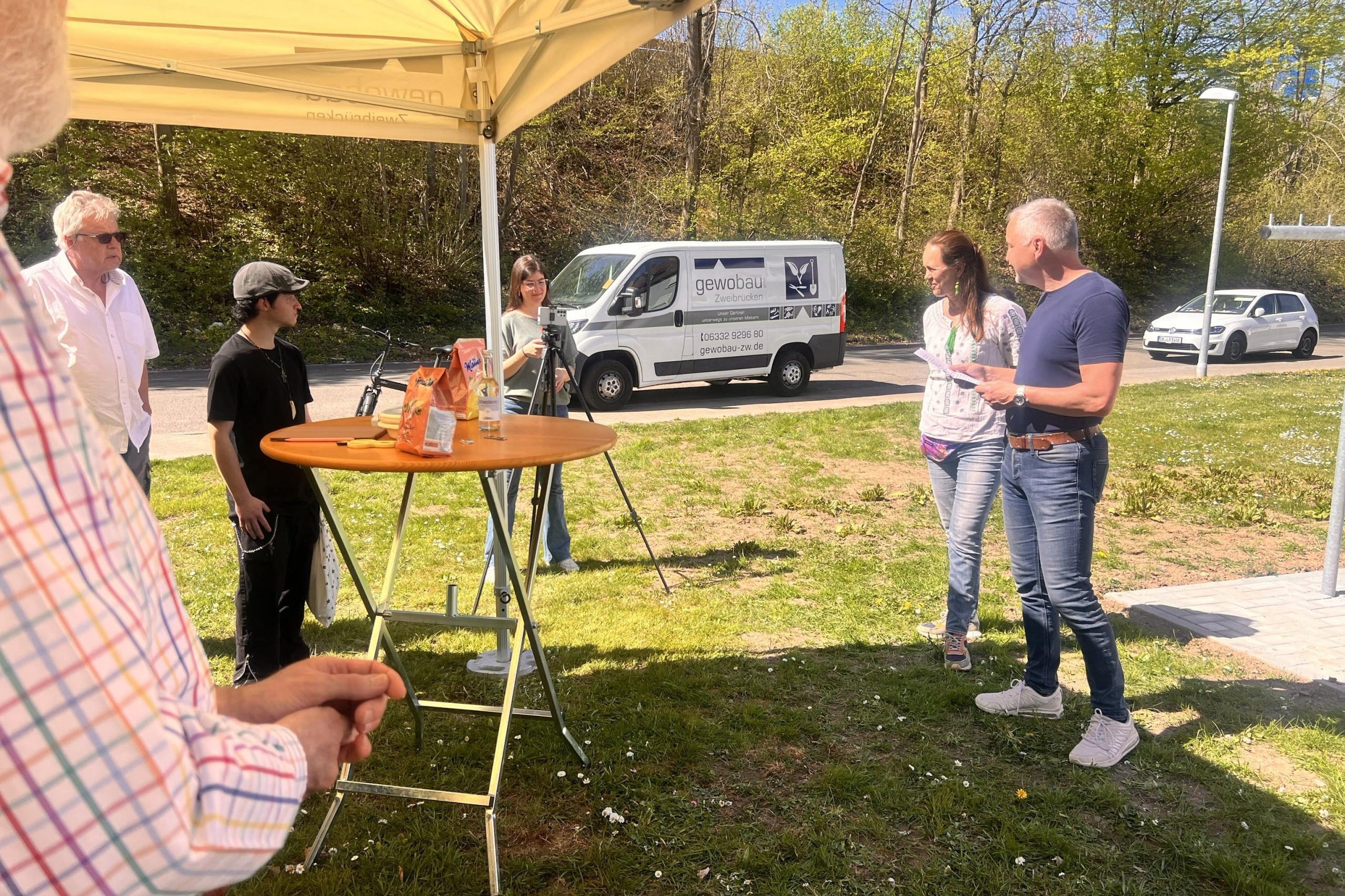 Infoveranstaltung auf einer Rasenfläche. Ein Redner im blauen T-Shirt. Eine Frau in weißer Bluse steht daneben. An einem Stehtisch sieht man Zuhörer