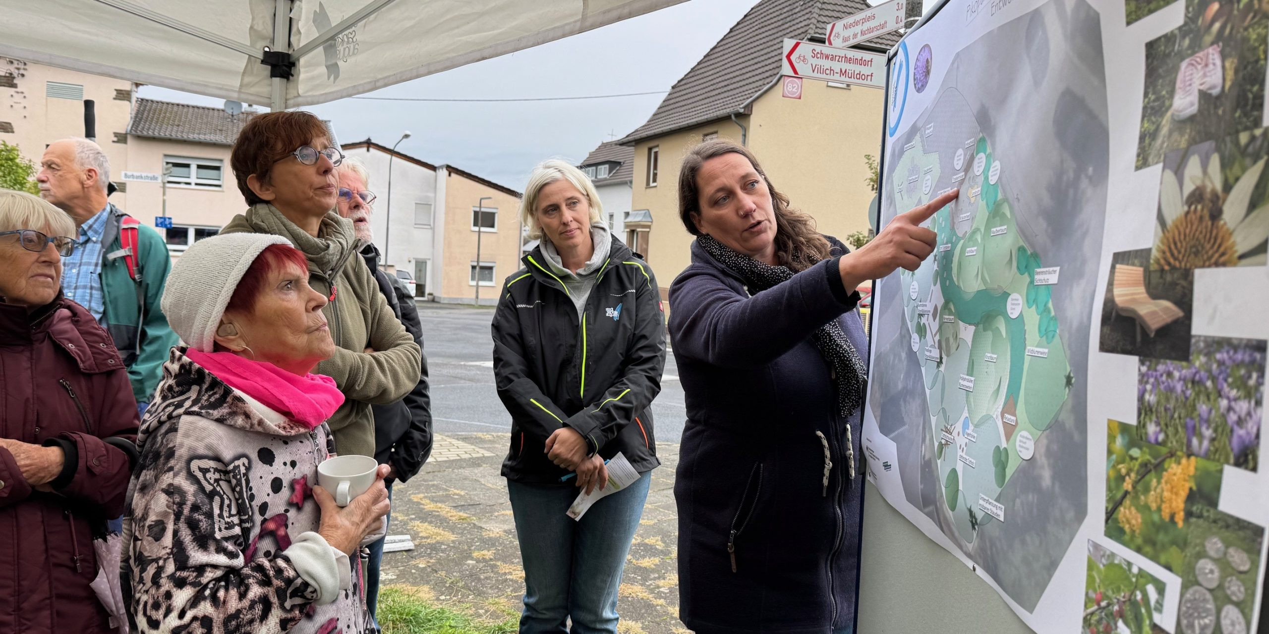 Eine Gruppe von Frauen und Männern steht vor einer Stellwand mit einem Gestaltungsplan. Eine Frau zeigt auf den Plan und erklärt etwas